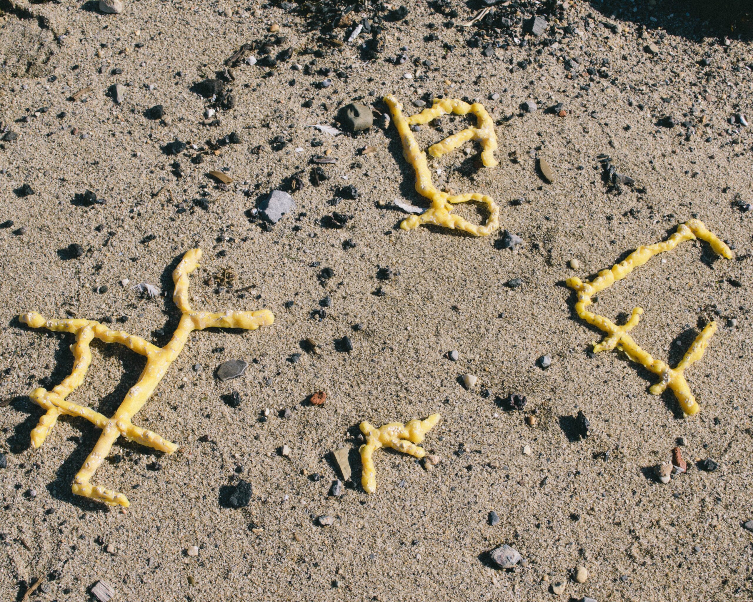 Three angular beeswax figures sit on rock-flecked sand.